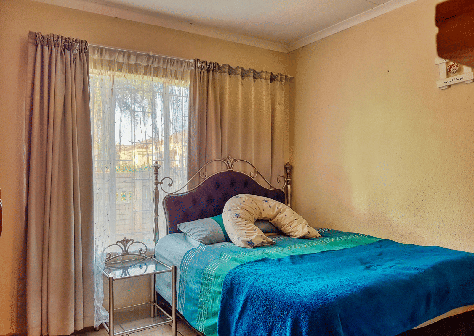 A bedroom featuring a double bed with blue sheets spread neatly over it with beige curtains covering the window behind it.
