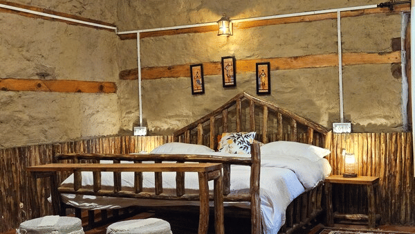 A close-up shot of the double bed at Hamta Peak Cottage featuring 2 round stools in front of the wooden bed frame in a brightly-lit room at Amara Upepo - The Sky Village, Manali.