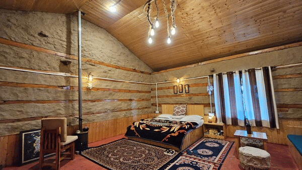Full-view shot of the Indrasan Peak Cottage showcasing a double bed surrounded by carpets on the floor along with a cosy coffee corner at Amara Upepo - The Sky Village, Manali.