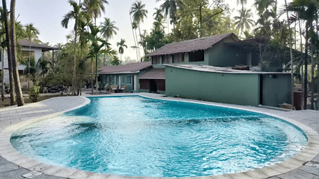 An outdoor swimming pool sits in front of a building surrounded by tall palm trees at Oceanature Resort, Alibaug.