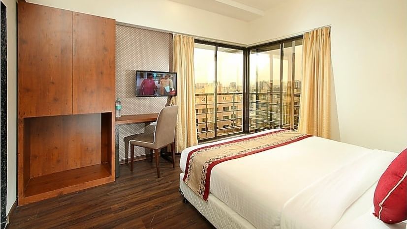 A bedroom featuring a wooden wardrobe, a study desk with a chair, a flat-screen TV, and large windows with curtains, offering an urban view.