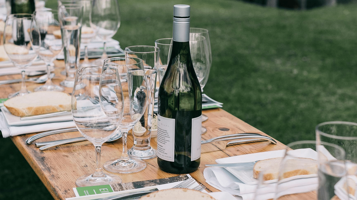 A long wooden table is set outdoors for a meal, featuring wine glasses, plates, cutlery, and a wine bottle.