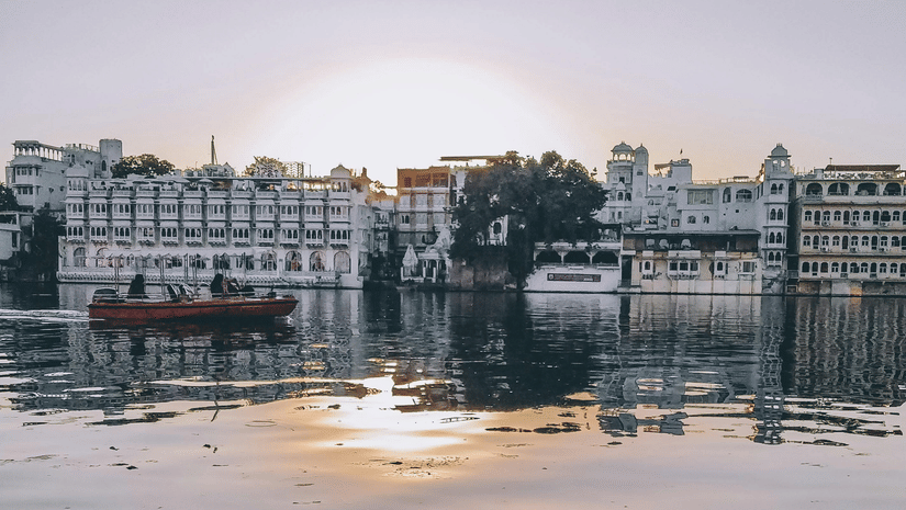 A beautiful shot of the Lake Pichola with calm waters overlooking several buildings with the sun setting behind it.