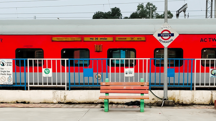 A train halted at Dehradun Railway Station separated by railings from a bench and pavement