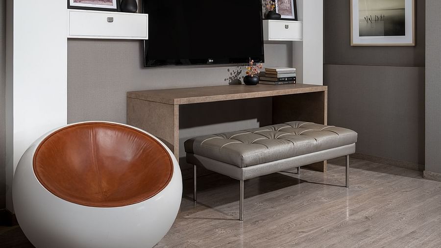 A modern room showcases a tall bookshelf, a wall-mounted television situated above a minimalist desk, and a stylish round upholstered chair, offering a comfortable space at The Atara, Gurugram