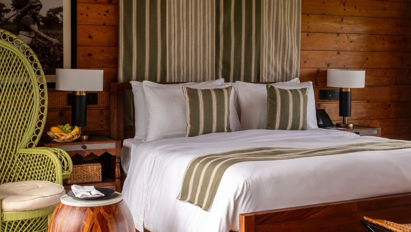 A vertical shot of a bedroom with a king-sized bed, a striped green and white canopy, and a large green wicker chair. A wooden side table and a large framed black and white photo on the wooden paneled wall complete the rustic, yet elegant look. This is a r