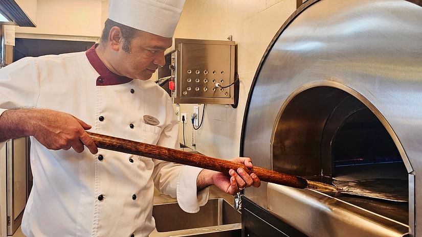 A chef in a white uniform carefully placing food into a traditional wood-fired oven in a professional kitchen