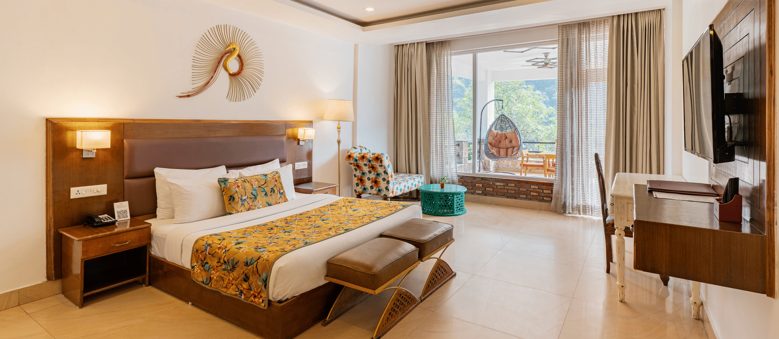A well-lit bedroom featuring a big bed facing a television screen, with an attached balcony featuring a standing swing chair overlooking the trees outside | Summit by the Ganges Beach Resort & Spa, Rishikesh