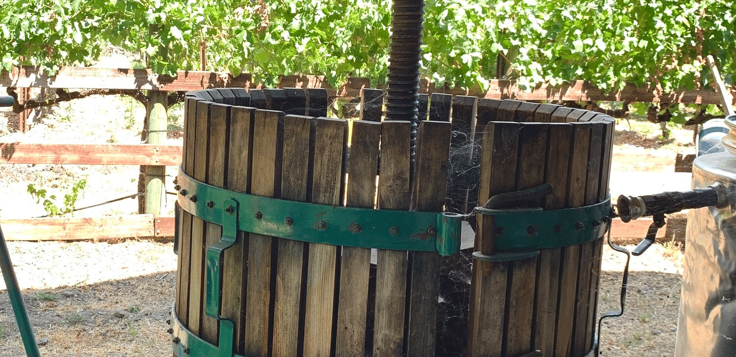A wooden barrel press with metal bands and a top mechanism stands on a platform with wheels, outdoors with greenery in the background.