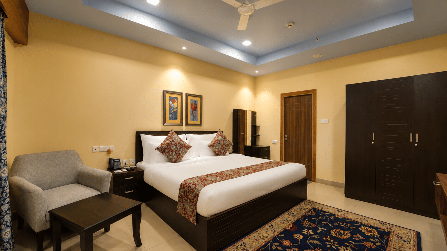 Deluxe suite bedroom with a king bed, grey armchair, coffee table, dark wooden wardrobes and a patterned rug at MAYFAIR World Cup Village, Rourkela.