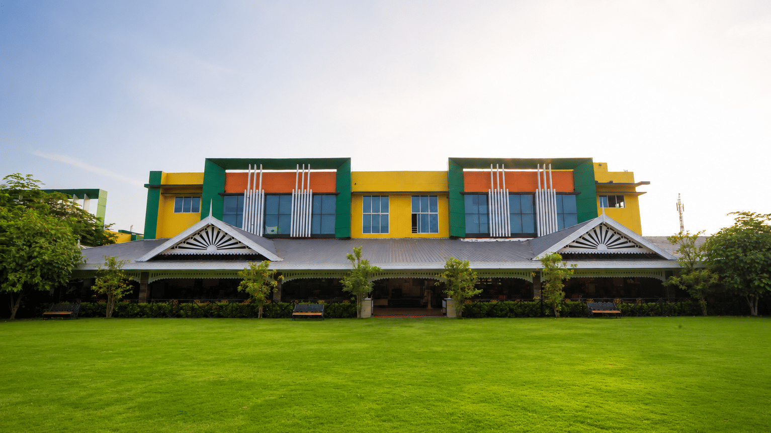 Vatika lawn with trimmed grass, garden benches and shrubs fronting a colourful building with a pitched roof at MAYFAIR World Cup Village, Rourkela.
