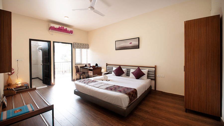 A brightly lit bedroom with a wooden floor, a large bed with purple bedding, an open suitcase on a bench