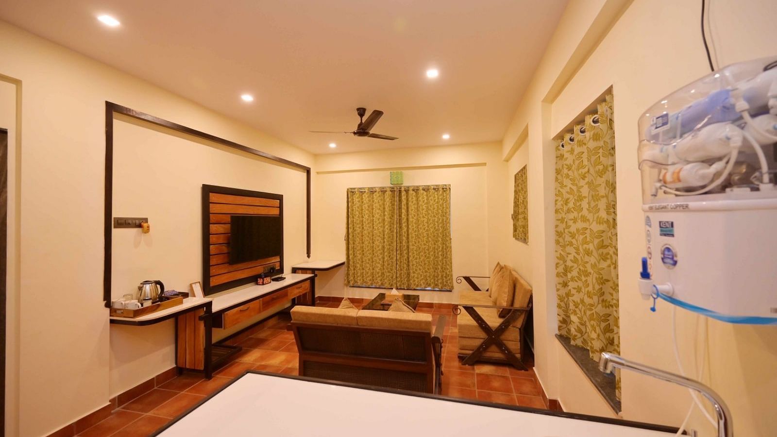 A living room with a TV mounted on a decorative wall panel, a couch, a ceiling fan, recessed lighting, and a water filter visible in the foreground.