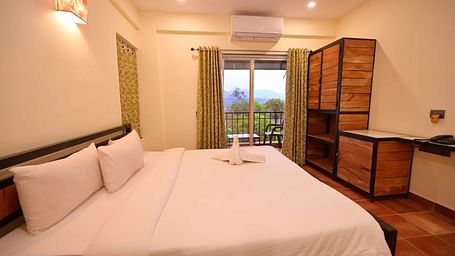 A bedroom with a dark wooden bed frame, white linens, a decorative wall lamp, an air conditioner, and a wooden cabinet, with a sliding glass door leading to a balcony.