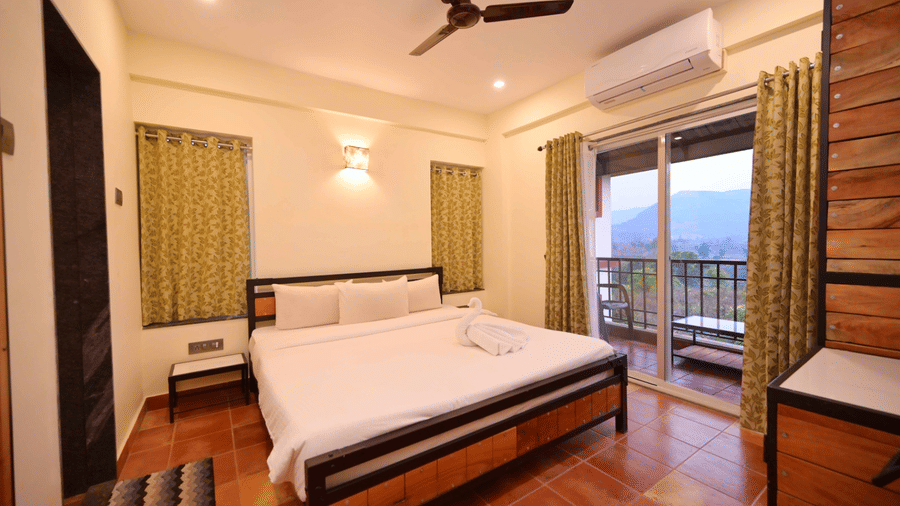 A bedroom with a wooden bed frame with white linens under a ceiling fan, an air conditioner over the sliding glass door leading to a balcony with a view