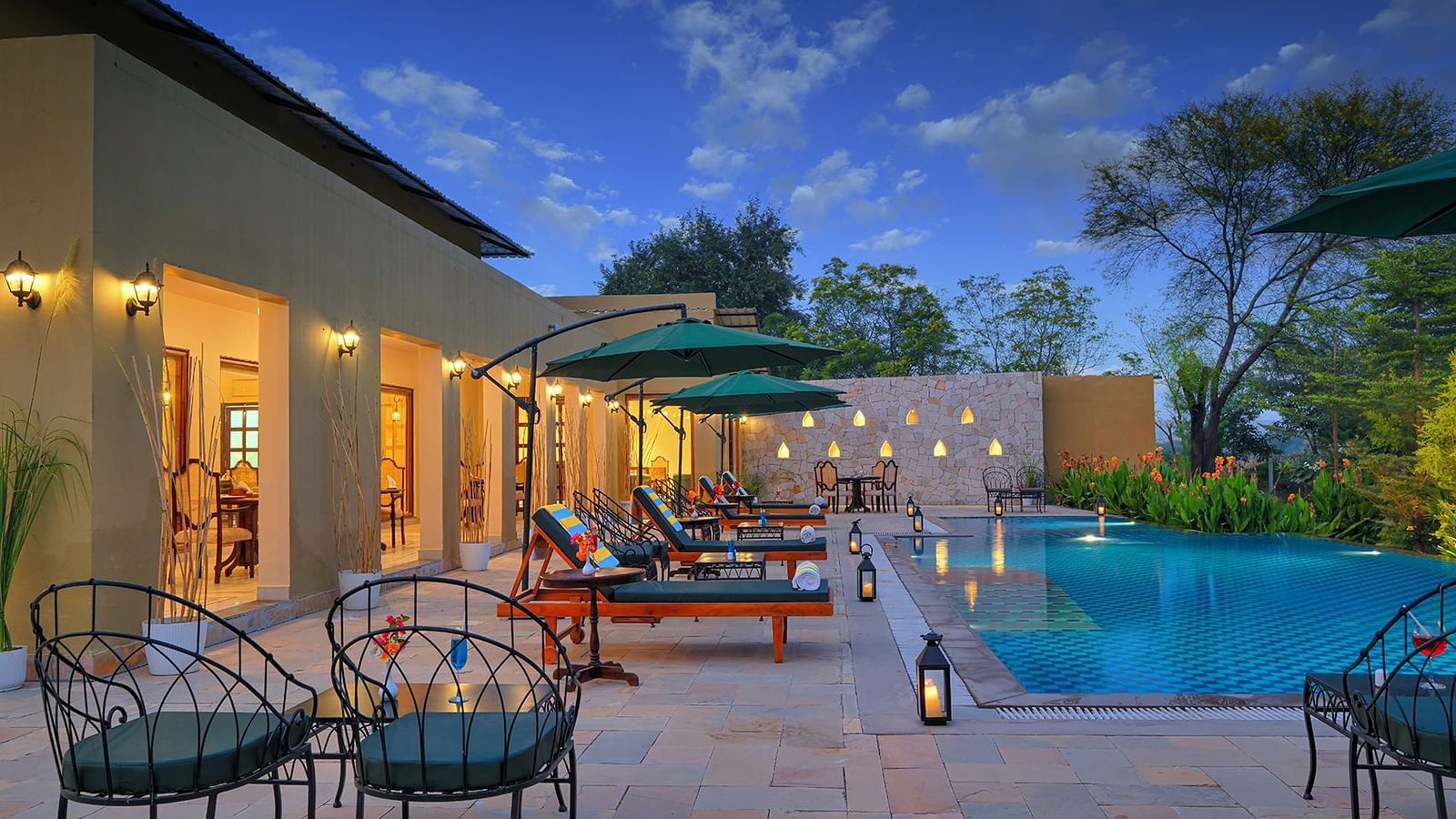 Outdoor patio with lounge chairs, umbrellas, and a sparkling blue swimming pool at dusk - Singhvilas, Ranthambore