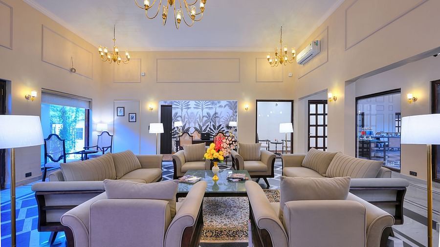 A spacious and elegantly decorated lounge area features multiple comfortable sofas and armchairs arranged around coffee tables, ample natural light from large windows, ornate chandeliers, and a polished floor with geometric patterns - Singhvilas, Ranthambo