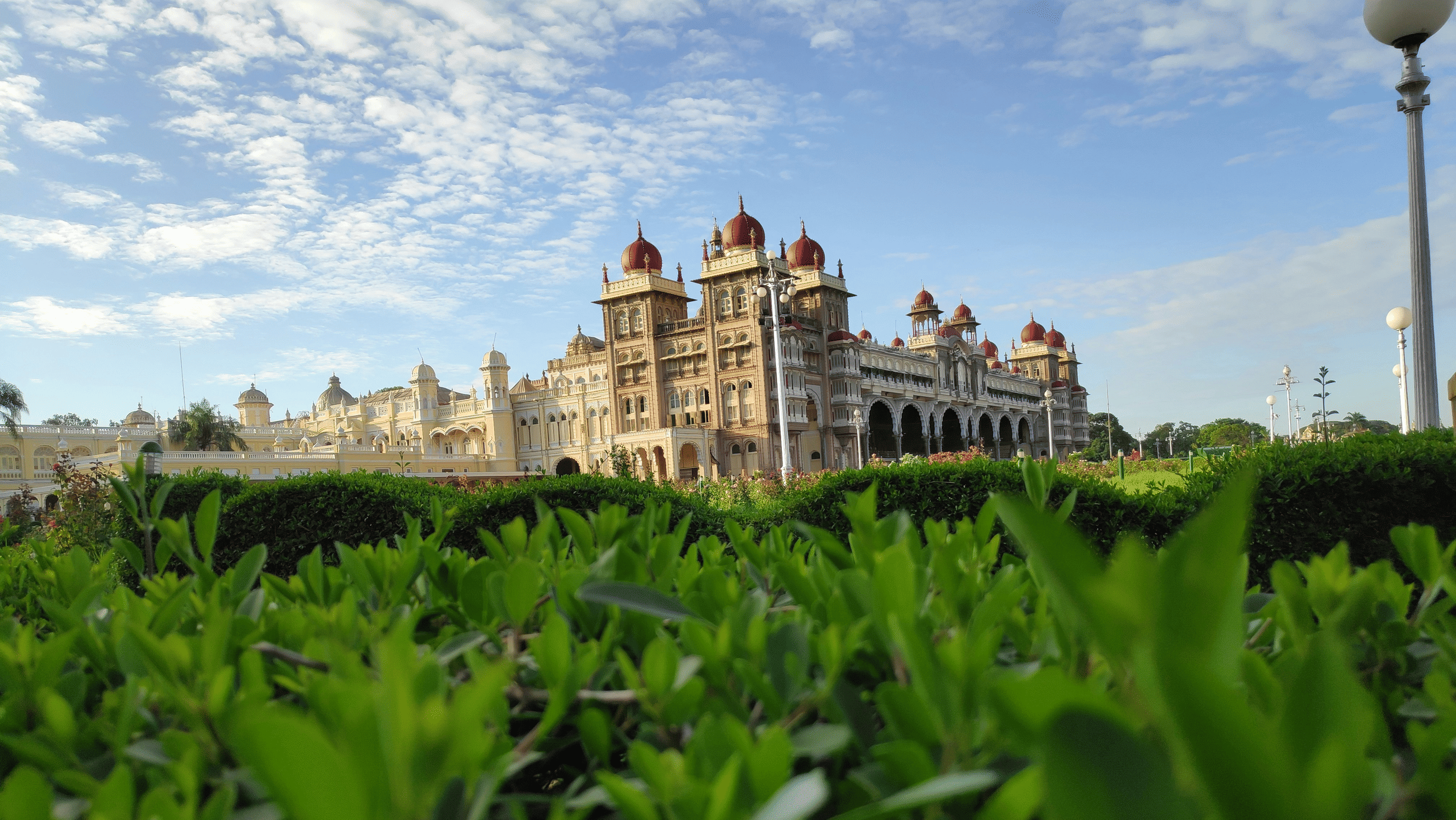 Daytime view of the Mysore Palace with its ornate domes and arches rising above lush green hedges and gardens under a bright blue sky.