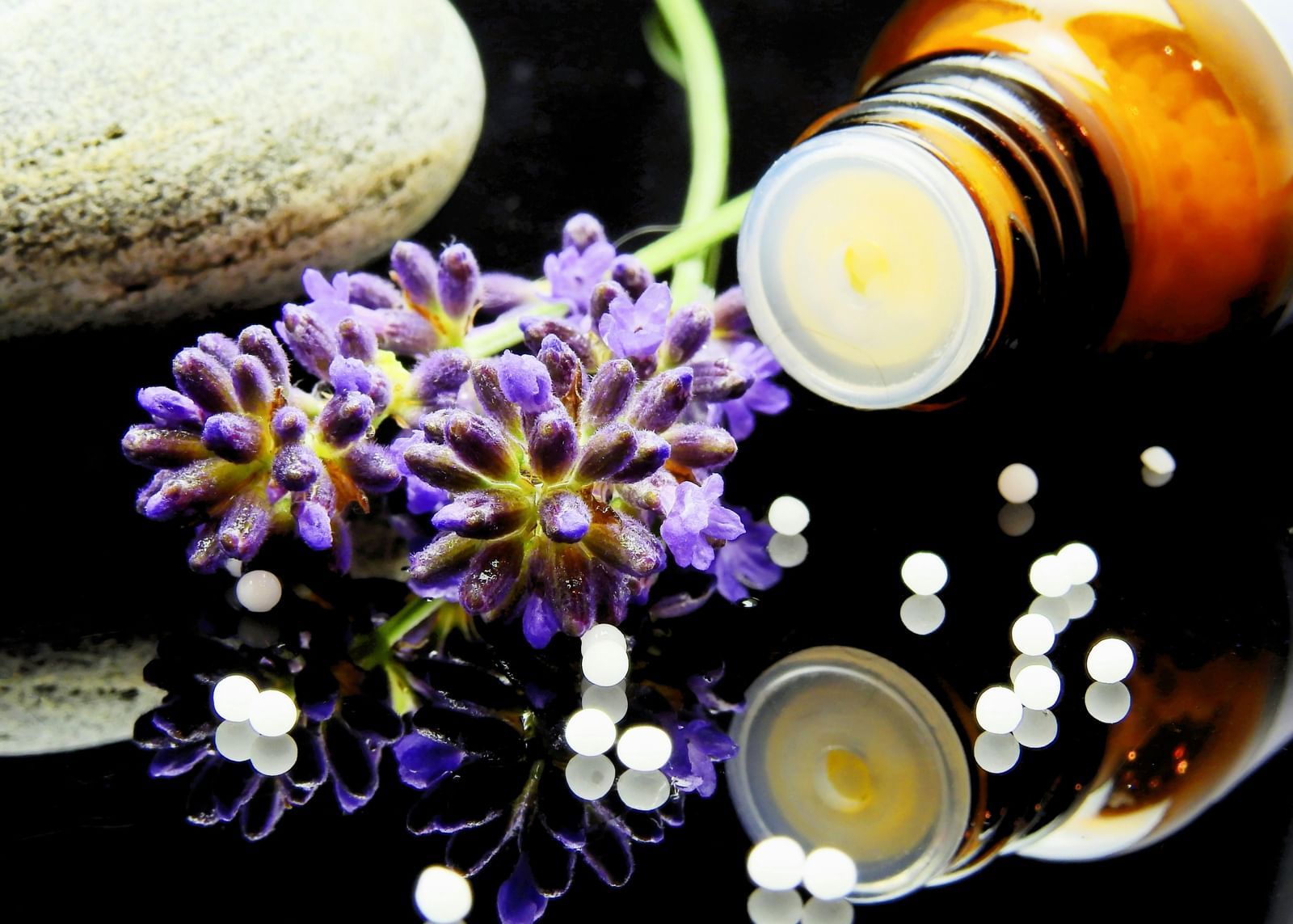 Homeopathic globules spilled from a brown glass bottle near lavender sprigs and a smooth stone on a reflective black surface.