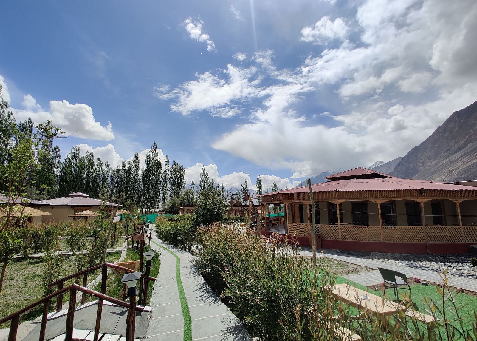 A scenic view of White Sands Resort, Ladakh featuring traditional buildings, lush greenery, and a clear sky with clouds.