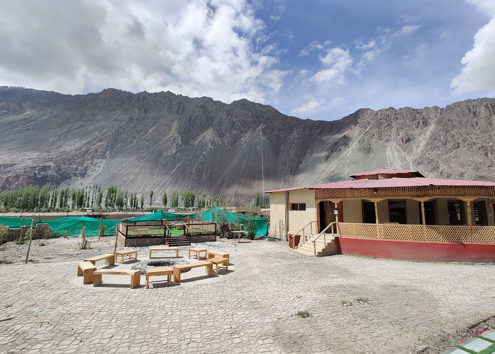 A rustic outdoor area with simple seating, set against a backdrop of rugged mountains and a cloudy sky at White Sands Resort, Ladakh.
