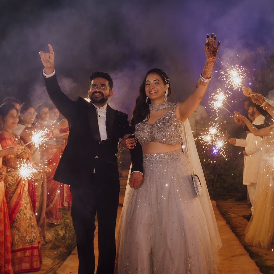 Bride and groom celebrating with arms raised, surrounded by guests and vibrant smoke effects at night.