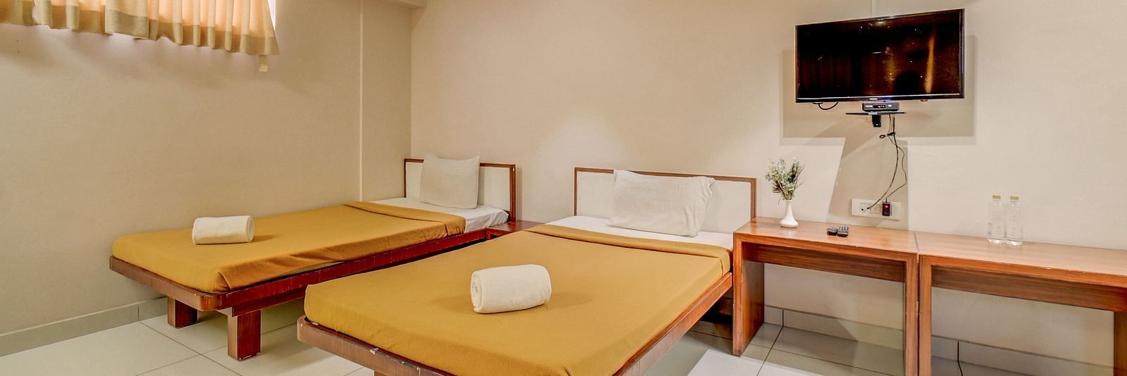 A spacious room at Harigovinda's JNStay, featuring a wall-mounted TV above a wooden table set against the wall, and two beds