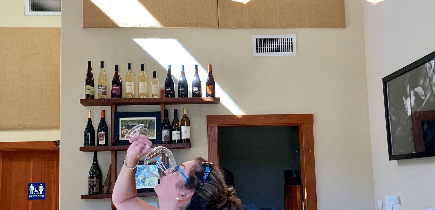 A women drinking a liquid from a glass with wine bottles placed on the wall.