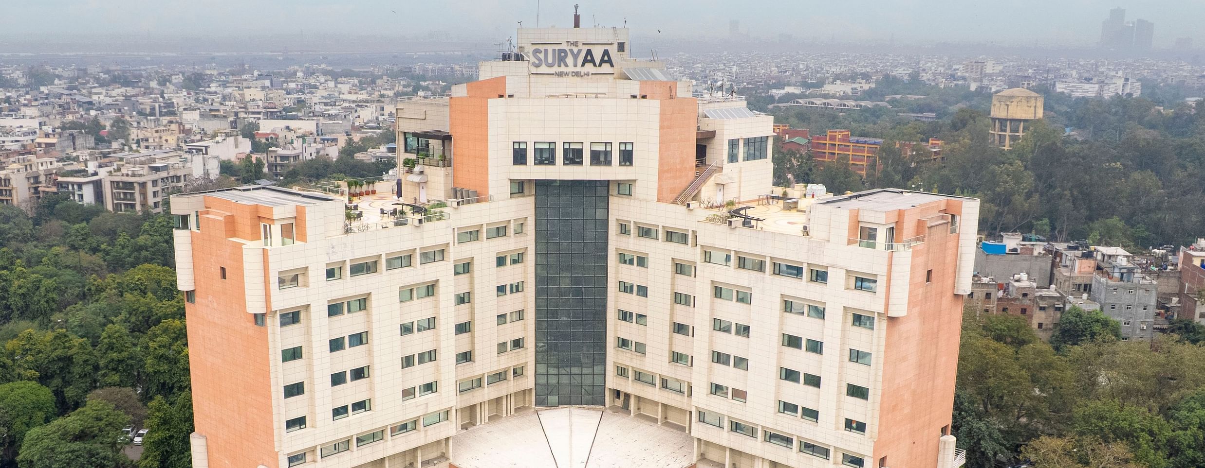 Aerial view of The Suryaa New Delhi showcasing the full hotel building, swimming pool, and surrounding cityscape.