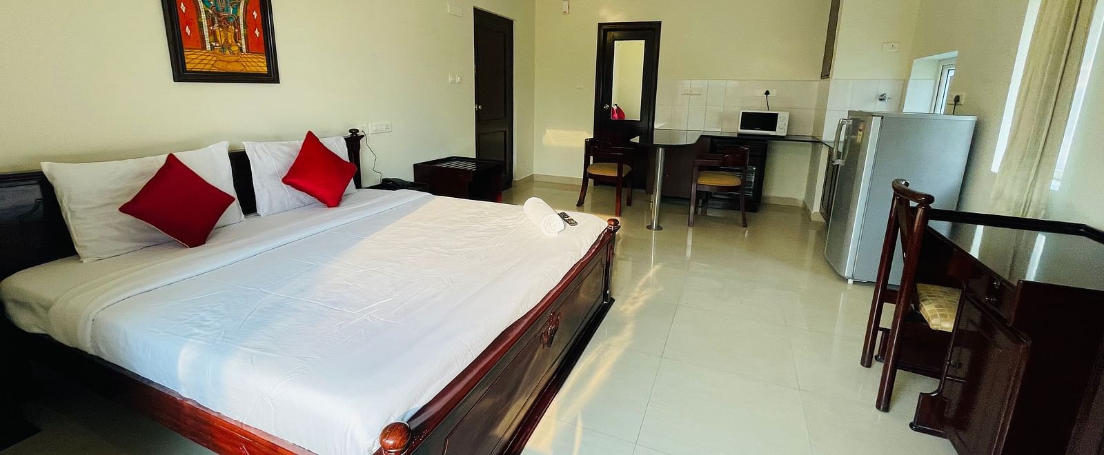 A room with a double bed featuring white linens and red cushions, a separate seating area, and a glimpse into a kitchenette space at Sanctum Suites Indiranagar, Bangalore.