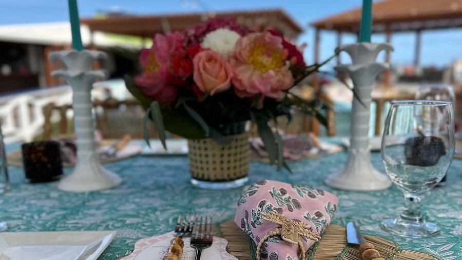 A dining table at the beachside featuring a bouquet the centre of the table, a table mat where knife and forks are placed, along with a wine glass and candle stands. 