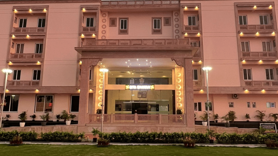 The front of a grand, light-pink building with an ornate entrance is illuminated in the evening - Kankidham Lords Inn