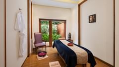 A serene spa room with a massage table, soft lighting, a chair, a robe hanging on the wall, and a window overlooking greenery - Ananta Spa and Resort, Pushkar.