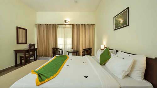 A bedroom with a neatly made-up bed includes green accents on the bed and curtains, along with a writing desk. - Abad Brookside Lakkidi, Wayanad