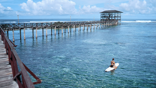 A long wooden pier extending over the sea with a person on a surfboard nearby at Lamari Siargao.