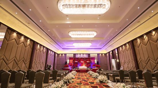 A view from the back of the banquet hall at Pride Premier Alwar, showing rows of grey upholstered chairs facing a decorated stage, set up for a formal event or conference.
