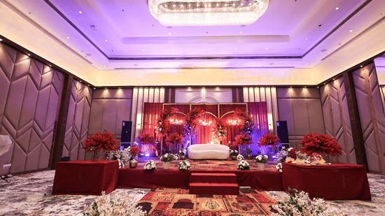A wide-angle view of a decorated banquet hall at Pride Premier Alwar, featuring a red and gold floral stage backdrop, tiered seating, and ornate wall paneling under warm recessed lighting.