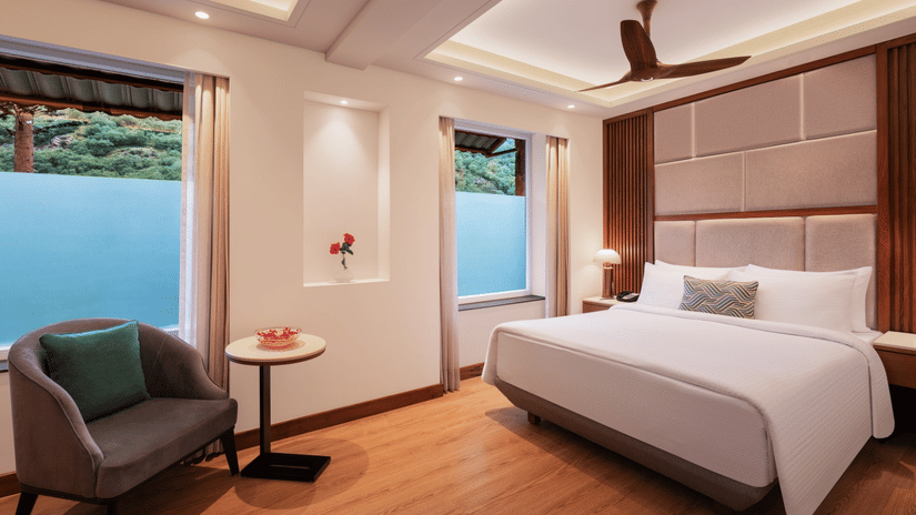 Deluxe Classic Room at Ananta Spa and Resort, Ajabgarh featuring a double size bed, a sofa chair, a centre table, ceiling fan, and windows.