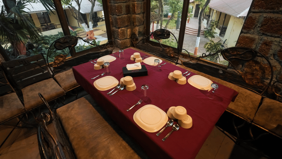 A long restaurant table dressed in a red tablecloth with clean plates, situated in a corner with large windows.