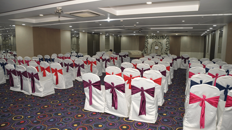 A spacious conference or wedding hall at Ambar Sarovar Portico, Gandhidham is arranged with multiple rows of chairs decorated with ribbons.