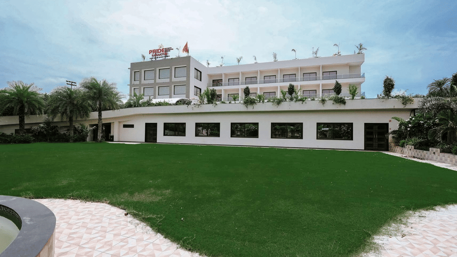 A wide shot of the Arawali Ananta Elite Jaipur hotel building with a green lawn in the foreground and a mountain in the background.