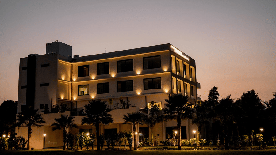 The hotel building of Arawali Ananta Elite Jaipur is illuminated at night with palm trees in front.