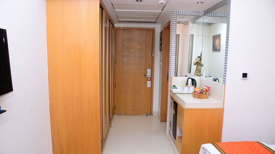 Hotel room entryway with wooden wardrobe, full-length mirror, tea and coffee station, snacks, and a glimpse of the bed with an orange throw - Citrus Classic, Outer Ring Road, Bangalore