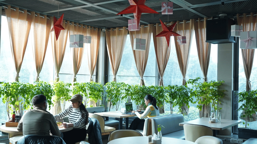 A covered open-air dining space of Falaknuma restaurant with round tables, lounge chairs, ceiling fans, decorative hanging elements, and guests seated near glass railings at Clarks Avadh, Lucknow.