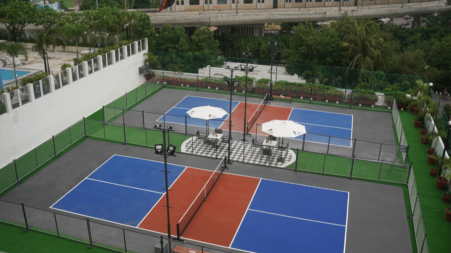 An elevated view of multiple fenced sports courts with blue and red surfaces, net partitions, surrounding greenery, and nearby buildings at Clarks Avadh, Lucknow.