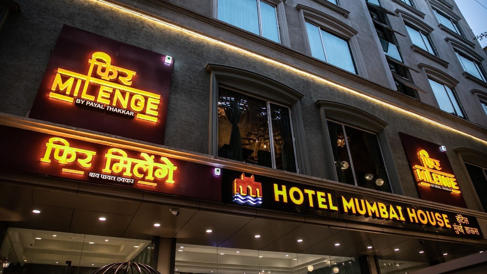 Exterior view of Hotel Mumbai House with illuminated signs for MILENGE BY PAYAL THAKKAR and HOTEL MUMBAI HOUSE in the evening.