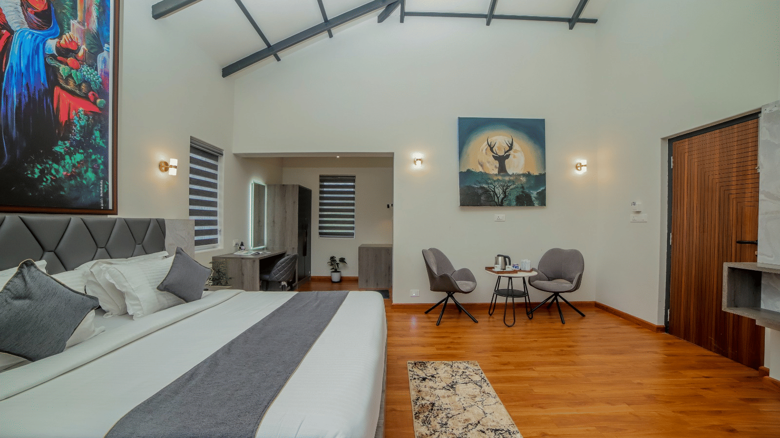 Side view of a bedroom in MYST Resorts, Doddabetta, Ooty, showcasing its seating area for 2, wooden flooring, a wardrobe, and a work desk.
