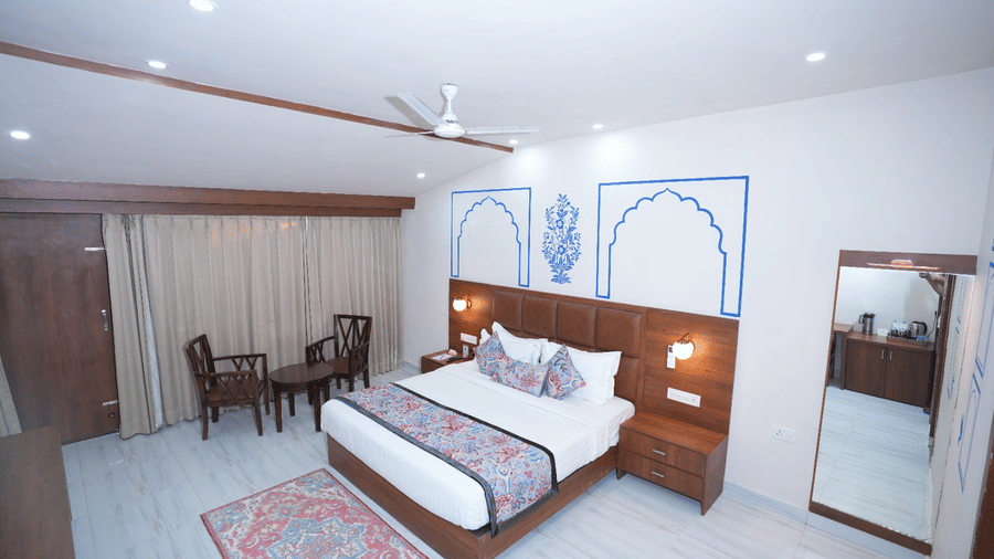 A spacious bedroom with a bed, curtains, wooden furniture, wall art, and a large mirror at Bamboo Saa Sunrise Wedding & Wellness Resort, Jaipur.