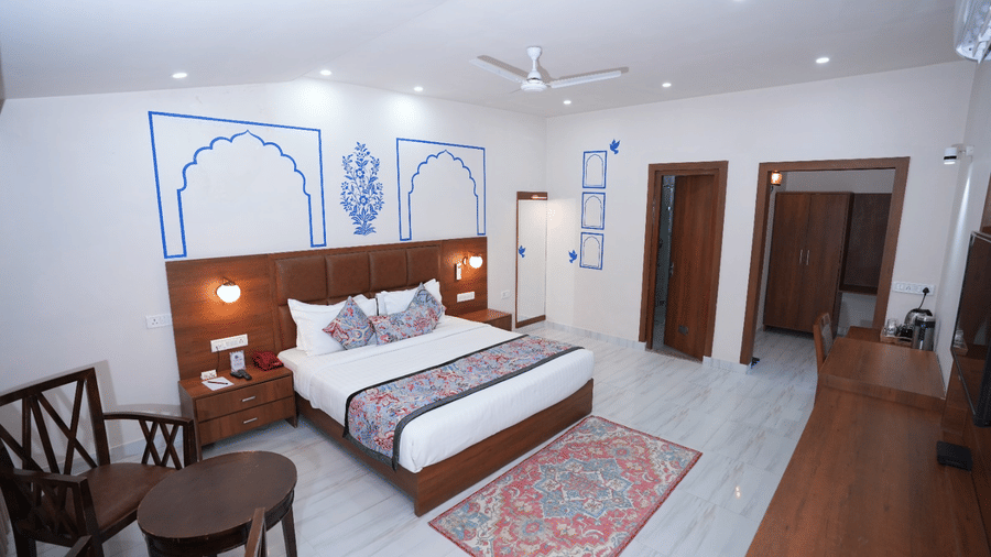 A bright bedroom featuring a double bed, wooden décor, seating, wall art, and a patterned rug at Bamboo Saa Sunrise Wedding & Wellness Resort, Jaipur.