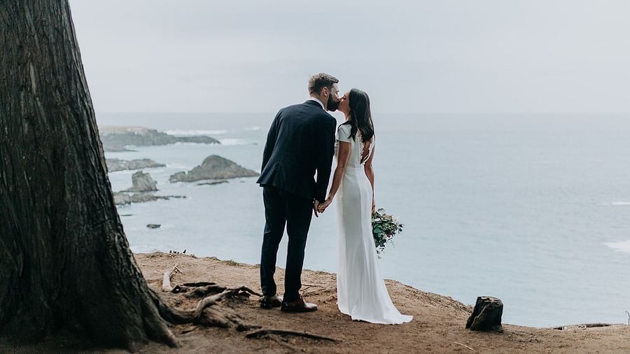 Bride and groom standing by a tree, overlooking the ocean
